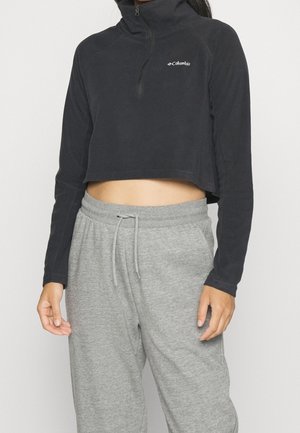 Vrouw draagt een zwarte korte Columbia fleece trui en grijze joggingbroek met trekkoord tegen een effen achtergrond.