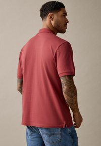 Camiseta polo marrón con mangas cortas y un cuello clásico, combinada con jeans azules. Presenta un corte recto y aberturas laterales en el dobladillo.