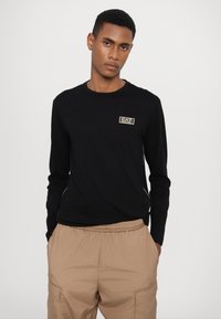 Camisa de manga longa preta com decote redondo e pequeno logotipo EA7 em dourado. Combinada com calças bege texturizadas de cintura elástica.