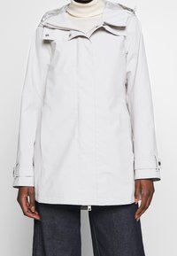 Světle šedá nepromokavá bunda se zipovým zapínáním, nastavitelnou kapucí a knoflíkovými manžetami. Disponuje bočními kapsami a hladkým, strukturovaným povrchem.