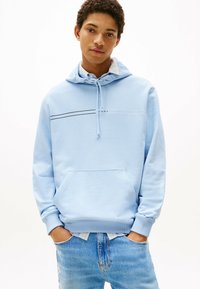 Felpa leggera blu con cappuccio, dotata di tasca a marsupio frontale, cappuccio con lacci e una striscia logata nera che attraversa il petto. Indossata sopra un paio di jeans blu sbiaditi.