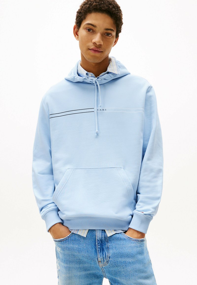 Felpa leggera blu con cappuccio, dotata di tasca a marsupio frontale, cappuccio con lacci e una striscia logata nera che attraversa il petto. Indossata sopra un paio di jeans blu sbiaditi.