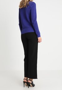 Stickad lila tröja med ett strukturerat mönster, tillsammans med svarta byxor med sidoknappar och svarta klacksandaler.