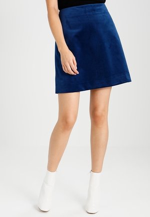 Jupe A-line en daim bleu avec une texture lisse, dotée d'une taille haute et d'un ourlet qui se termine au-dessus du genou, associée à des bottines blanches à talons.