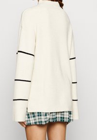 Suéter crema con textura acanalada, cuello alto y rayas horizontales negras en las mangas; usado sobre una falda de cuadros verdes.