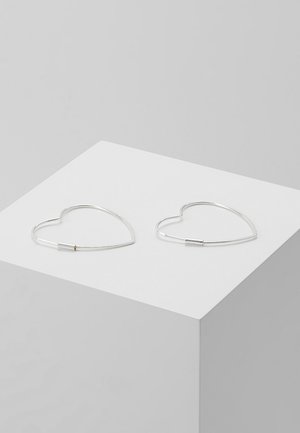 Coppia di orecchini a cerchio a forma di cuore in filo d'argento posati su un cubo bianco su sfondo grigio chiaro.