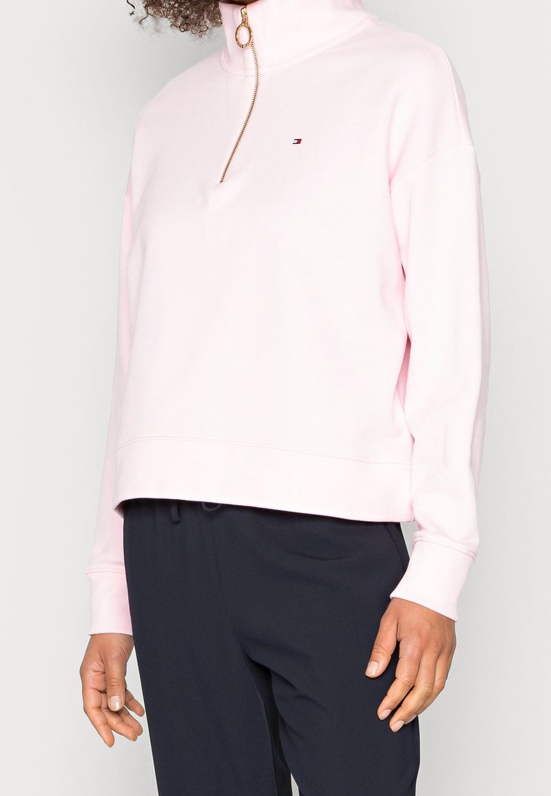 Sudadera de color rosa con cuello alto y cremallera dorada. Presenta un pequeño logo en el pecho y hombros caídos. Tejido suave y corte relajado.