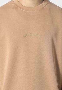 Béžová mikina s kulatým výstřihem a krátkými rukávy, s vyšitým nápisem "PAS DE MONACO" zlatou nití přes hruď.