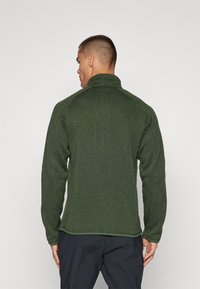 Green fleece jacket with a high collar, long sleeves, textured fabric, and a fitted design. Back view shows smooth, minimal detailing.