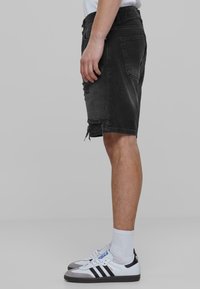 Shorts de mezclilla negra con dobladillo desgastado, bolsillos y un ajuste relajado. Combinados con zapatillas blancas y negras y calcetines blancos.