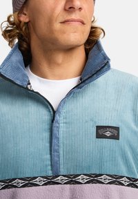 Pullover in velluto a coste azzurro chiaro con colletto zip, caratterizzato da una toppa logo nera e una fascia decorata in grigio e bianco nella parte inferiore.