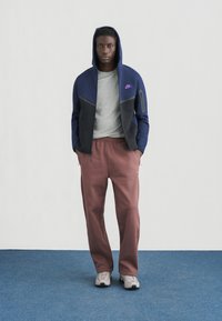 Nike Kapuzenjacke mit Reißverschluss in Marineblau und Schwarz mit grauem Innenfutter, kombiniert mit kastanienbraunen Jogginghosen und hellgrauen Sneakers, stehend auf blauem Boden.