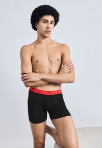 Μαύρα βαμβακερά μπόξερ με κόκκινη ζώνη μέσης που έχει το λογότυπο "TOMMY HILFIGER". Το μοντέλο έχει φυσικό χτένισμα και σταυρωμένα χέρια.