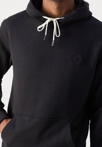 Sudadera de algodón negra con un bolsillo frontal, cordones ajustables y un pequeño logo negro en el pecho. Textura suave y diseño casual.