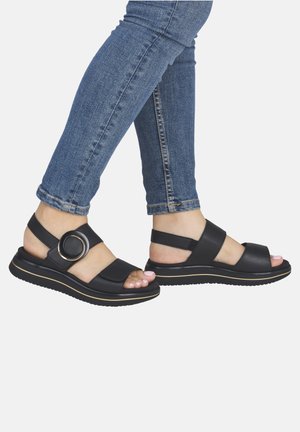 Benen met een blauwe spijkerbroek en zwarte open-teen sandalen met een dikke zool en een grote gesp rond de enkel.