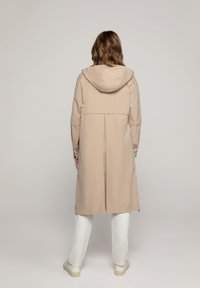 Beige waterdichte jas met een capuchon, voorzien van een plooi aan de achterkant, verstelbare mouwen en een middellange snit. Ggedragen over witte broek.
