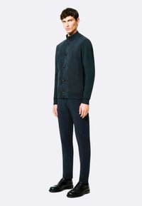 Giubbotto bomber in suede nero con colletto alto, bottoni frontali e polsini a coste, abbinato a pantaloni scuri e scarpe stringate nere.