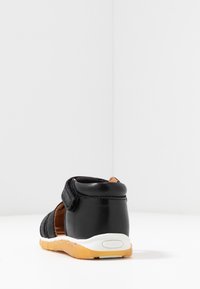 Černá kožená bota s suchým zipem, nízký profil, texturovaný svršek, polstrovaný límec a gumová podrážka s béžovým akcentem.