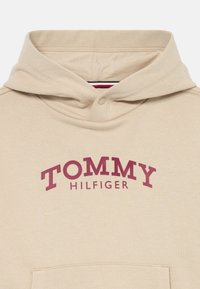 Μπεζ φούτερ με κουκούλα και κουμπί πίεσης, με τη γραφή "TOMMY HILFIGER" σε έντονα κόκκινα γράμματα και μπροστινή τσέπη.