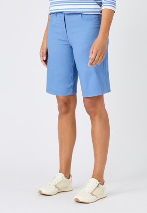 Persona que lleva pantalones cortos azul claro hasta la rodilla, camiseta de rayas y zapatillas blancas, de pie sobre un fondo blanco liso.