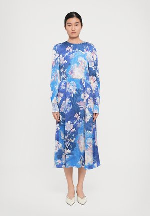 Woman standing in a blue floral long-sleeve midi dress paired with white pointed heels against a plain white background.