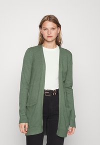 Groene gebreide cardigan met een relaxte pasvorm, open voorkant, lange mouwen en twee voorzakken, gecombineerd met een wit t-shirt en zwarte broek.