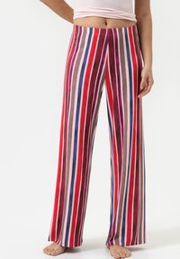 Un pantalon large en matière douce avec des rayures verticales rouge, rose, violet et marron. Taille élastique ; détail de pieds nus visible.
