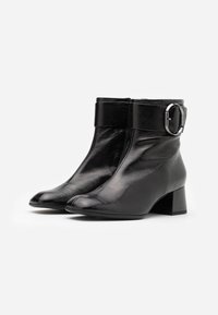 Botas de tobillo de cuero negro con punta cuadrada, acabado brillante, tacón medio y una correa ancha con una hebilla metálica redonda.