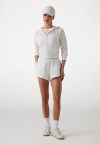 Bijela zip-up dukserica s logotipom, odgovarajuće bijele šortse, bijele tenisice i bijela kapa. Glatka tkanina, ležeran kroj i kratki rukavi.
