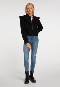 Giacca nera con zip e colletto arricciato, aderente in vita. Indossata con jeans skinny blu strappati e stivali neri con lacci.