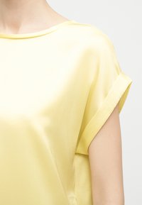 Μαλακό κίτρινο σατέν μπλουζάκι με κοντά, γυρισμένα μανίκια και φαρδιά λαιμόκοψη σε ανοιχτό φόντο.