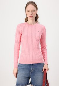 Rosa strikket genser med kabelmønster, rund hals, lange ermer og ribbestrikket kant. Brukt med denimshorts og en håndveske.