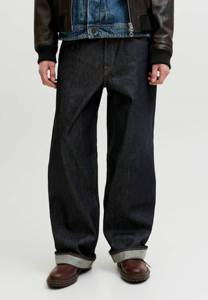 Personne portant un jean large et foncé retroussé, des bottes en cuir marron, une veste en jean sous une veste en cuir noire, debout sur un fond uni.