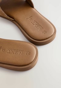 Brown slip-on sandals featuring a smooth leather upper, cushioned footbed, and horizontal stitching on the sole. Brand logo embossed on the footbed.