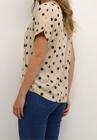 Haut beige à manches courtes avec des pois noirs, offrant une coupe ample et un ourlet arrondi. Associé à un jean bleu foncé.