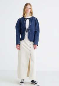 En mørkeblå denimjakke med puffede ærmer og knytelukning, kombinert med en kremfarget midiskjørt med frontknapper og splitt.