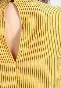 Chemise jaune à fines rayures verticales noires, avec un col en V et une fermeture à boutons au niveau du col. Le tissu semble lisse et léger.