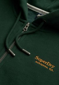 Sudadera con cremallera en verde oscuro, con cordones, cremallera metálica y bordado naranja que dice "SuperDry Athletics, CA." en el pecho.