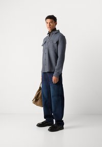 Blue herringbone shirt with chest pockets, paired with loose dark denim jeans and black shoes. Model holds a brown shoulder bag.