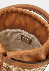 Brun faux læder spandtaske med samlet top, beige foring og 'GUESS'-logo tag. Har et vævet design på ydersiden.