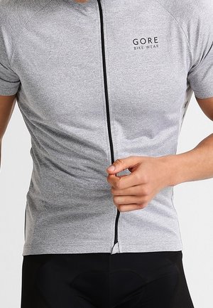 Muž nosí šedý krátký cyklistický dres na zip s logem "GORE BIKE WEAR" na hrudi a upravuje zip.