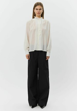 Witte blouse met lange mouwen, een gerimpelde voorkant, knoopsluiting en een hoge kraag, gecombineerd met zwarte broek met wijde pijpen en puntige schoenen.