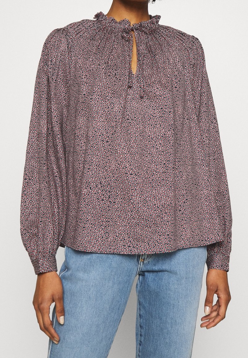 Blouse à manches longues en tissu à motifs avec un col froncé, une ouverture en chasse et des poignets élastiques, présentée avec un jean délavé.