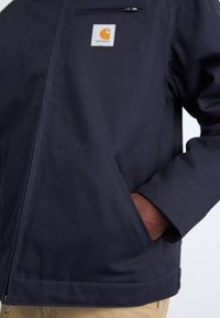 Veste bleu marine en tissu résistant avec une poche zippée sur la poitrine et des poches latérales ; présente un patch logo Carhartt sur la poitrine gauche.