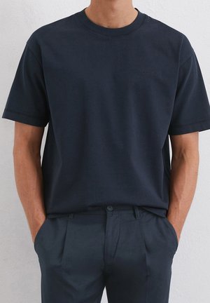 Mand iført en ensfarvet mørk marineblå t-shirt med korte ærmer og rund hals, stoppet ned i matchende mørk marineblå bukser med hænderne i lommerne.