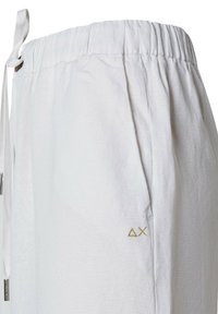 Pantaloni bianchi in lino con vita elastica, tasche laterali e un accento in logo triangolare dorato. Tessuto leggero e testurizzato.