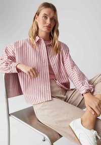 Jasnoróżowa koszula w paski na guziki z opuszczonymi ramionami, zestawiona z beżowymi spodniami i białymi butami niskimi, siedząca na metalowym krześle.