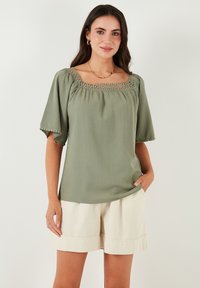 Light olive green top with a square neckline, lace detailing along the edge, and short sleeves. Paired with beige shorts.
