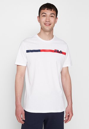 Jeune homme portant un t-shirt blanc FILA avec une bande rouge et marine, debout devant un fond gris clair uni, souriant et regardant devant lui.