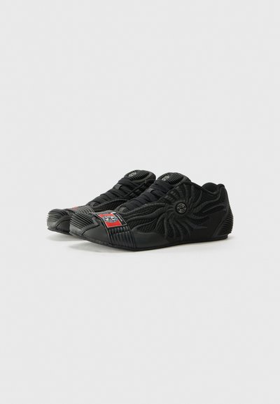 Des chaussures de sport noires présentent un dessus en mesh texturé, des accents en caoutchouc, un design de coque distinctif à l'avant et un logo bien visible sur le côté.